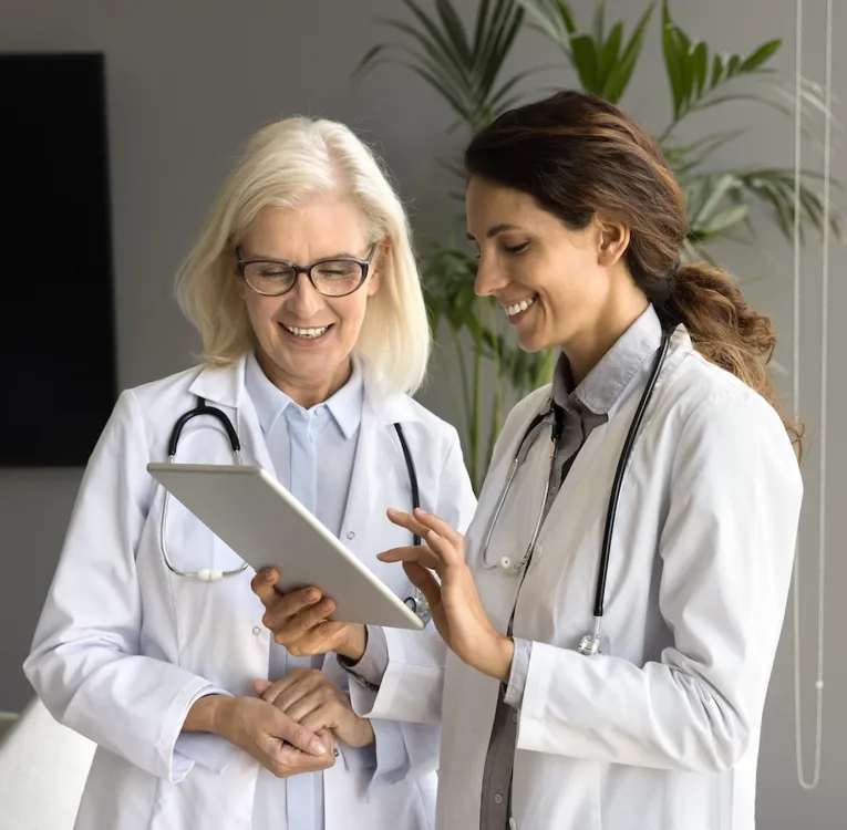 Två kvinnliga läkare i vita rockar med stetoskop granskar medicinsk information på en surfplatta i ett modernt kontor med stora fönster.