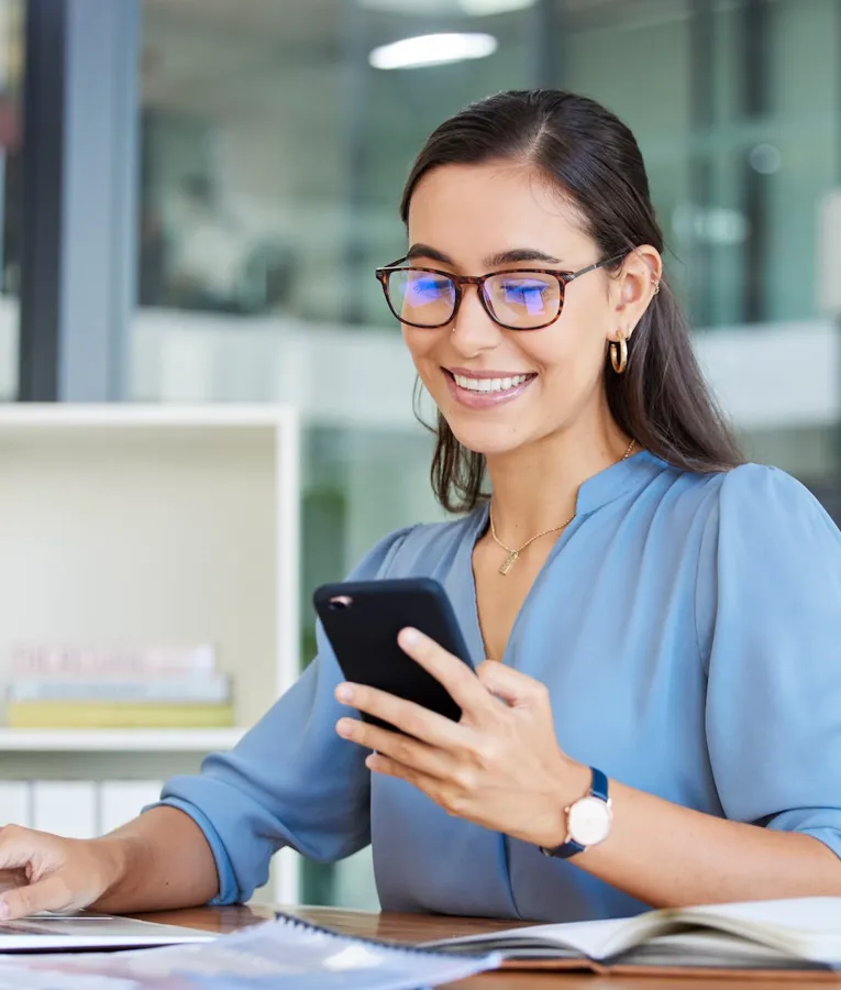 Lächelnde Büroangestellte in blauer Bluse nutzt Smartphone und Laptop am Schreibtisch, ein Symbol für digitale Kommunikation, hybrides Arbeiten und moderne Produktivität.
