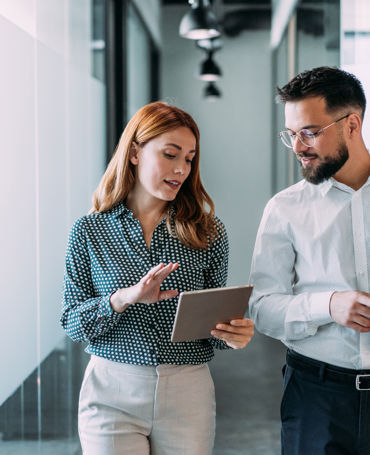 Zwei Mitarbeitende gehen durch das Büro und arbeiten gemeinsam an einem Tablet – ein Symbol für datengestütztes Workforce Management, moderne HR-Prozesse und effiziente Personalplanung.