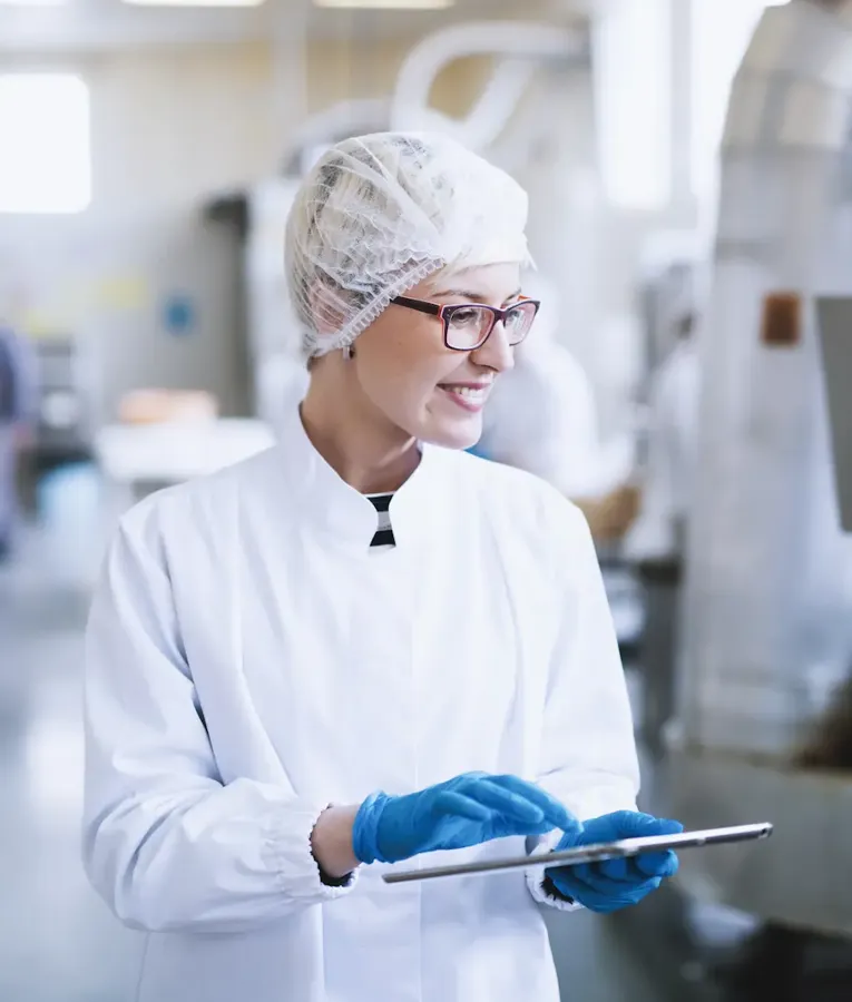 Eine lächelnde Mitarbeiterin in einem Lebensmittelverarbeitungsbetrieb nutzt ein Tablet zur Qualitätskontrolle und zur Steuerung der Arbeitsabläufe. Symbolisiert digitale Lösungen in der Lebensmittelproduktion.