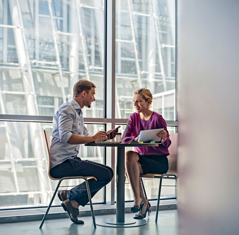 Ein Mann und eine Frau führen ein Geschäftsgespräch an einem kleinen runden Tisch in einem modernen Büro mit großen Glasfenstern. Der Mann hält ein Smartphone, während die Frau in einer lila Bluse ein Tablet hält und lächelt.
