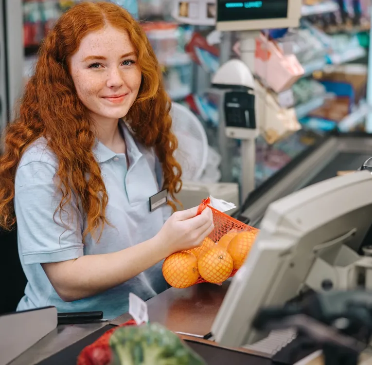 Lächelnde junge Kassiererin scannt Orangen im Supermarkt – Sinnbild für effiziente Personalplanung, moderne Kassensysteme und exzellenten Kundenservice.