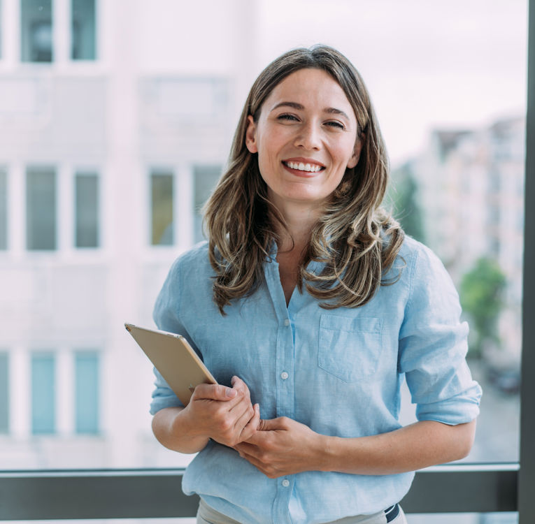 Eine lächelnde junge Frau mit langen braunen Haaren und hellblauer Bluse steht in einem modernen Büro und hält ein Tablet in der Hand. Sie strahlt Selbstbewusstsein aus – im Hintergrund große Fenster mit Blick auf die Stadt.