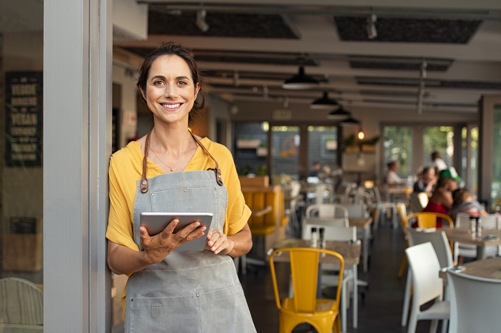 Eine lächelnde Restaurantinhaberin oder Managerin in gelber Bluse und grauer Schürze steht mit einem Tablet in der Hand am Eingang eines gut besuchten Cafés. Sie wirkt selbstbewusst und einladend, während sie den täglichen Betrieb und den Kundenservice koordiniert.