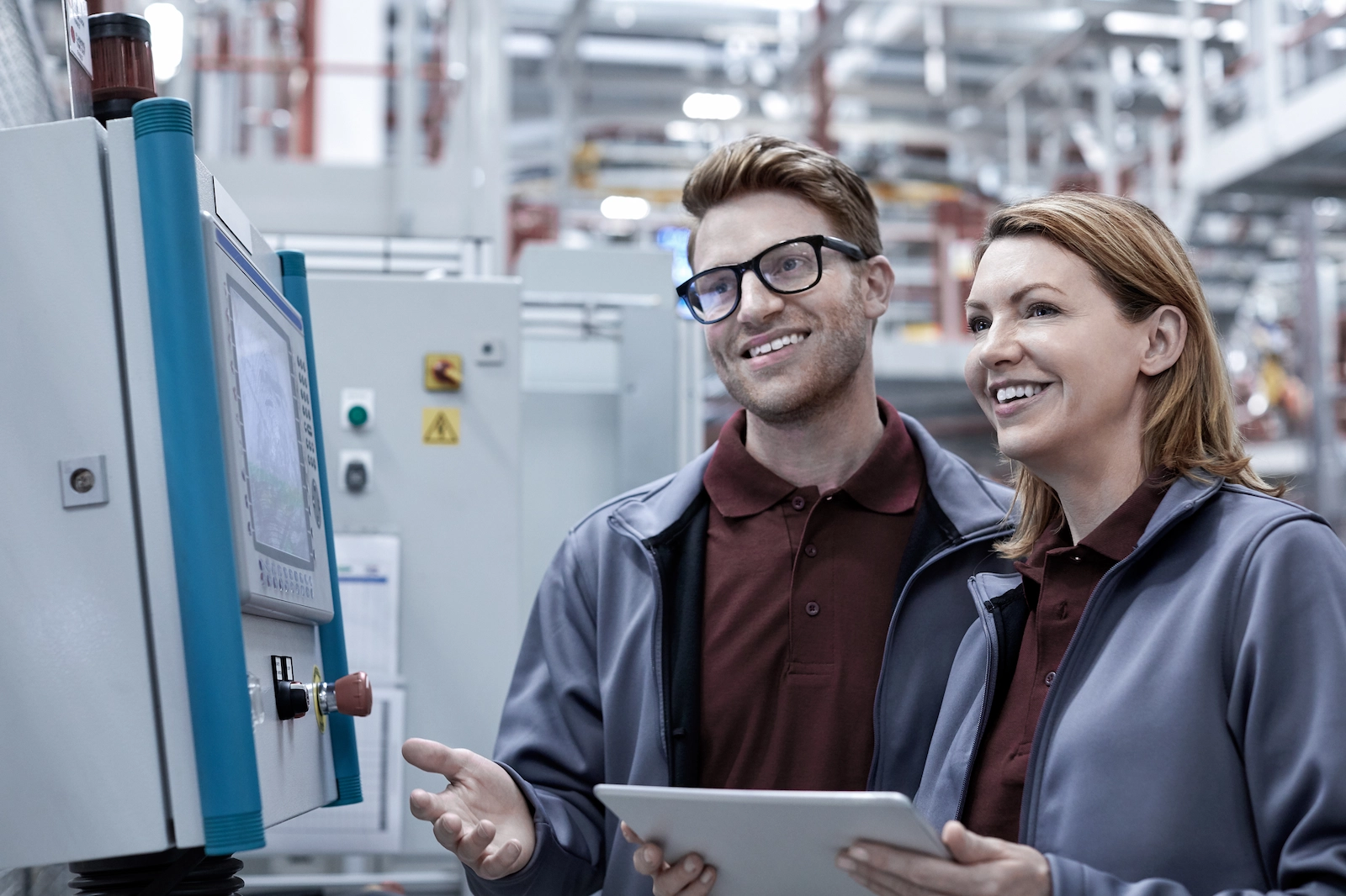 Zwei lächelnde Fabrikmitarbeitende nutzen ein Tablet und überwachen ein Kontrollpanel in einer modernen Produktionsanlage, was für Automatisierung und industrielle Effizienz steht.