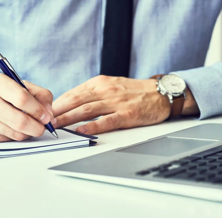 Nahaufnahme eines Geschäftsmannes in blauem Hemd und Krawatte, der Notizen in ein Notizbuch schreibt, während er an einem Laptop an einem modernen Schreibtisch arbeitet.