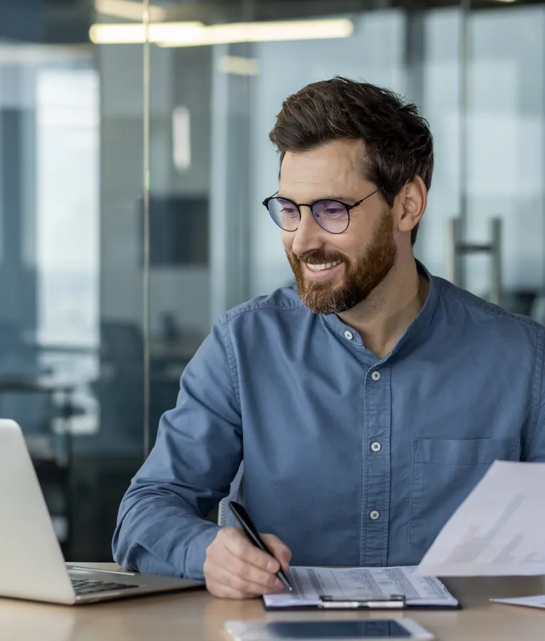 Ein lächelnder Geschäftsmann analysiert Diagramme und Berichte an seinem Schreibtisch und nutzt dabei ein Laptop zur Einsatzplanung und datenbasierten Entscheidungsfindung. Symbolisiert effizientes HR-Management und eine effektive Unternehmenssteuerung.