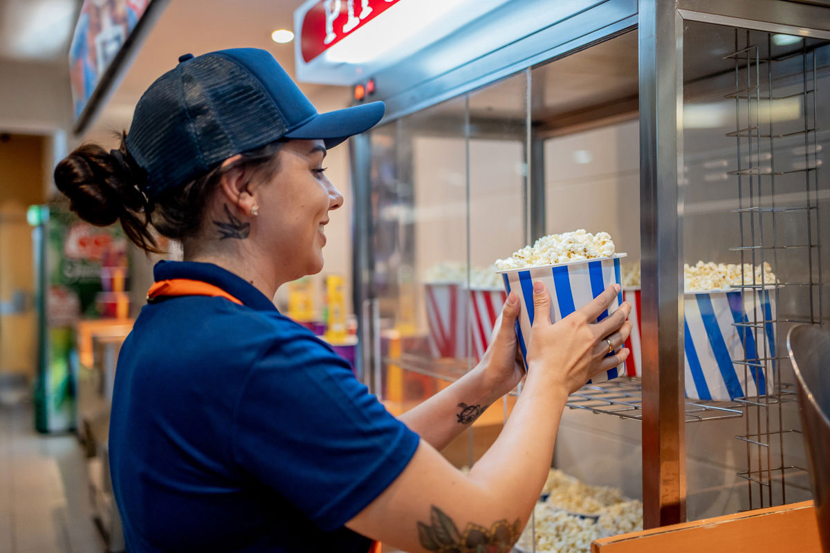 Lächelnder Kinomitarbeiter in Uniform stellt einen gestreiften Popcorneimer in einen Wärmeschrank, ein Symbol für Schichtarbeit, Einzelhandelsservice und effizientes Frontpersonal.