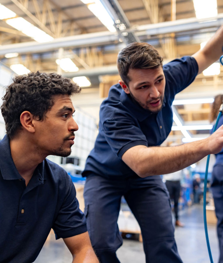 Zwei Techniker in marineblauen Uniformen arbeiten gemeinsam an der Wartung von Anlagen in einer Fabrik, mit Fokus auf Fachkräfte, Schulung und technische Abläufe.