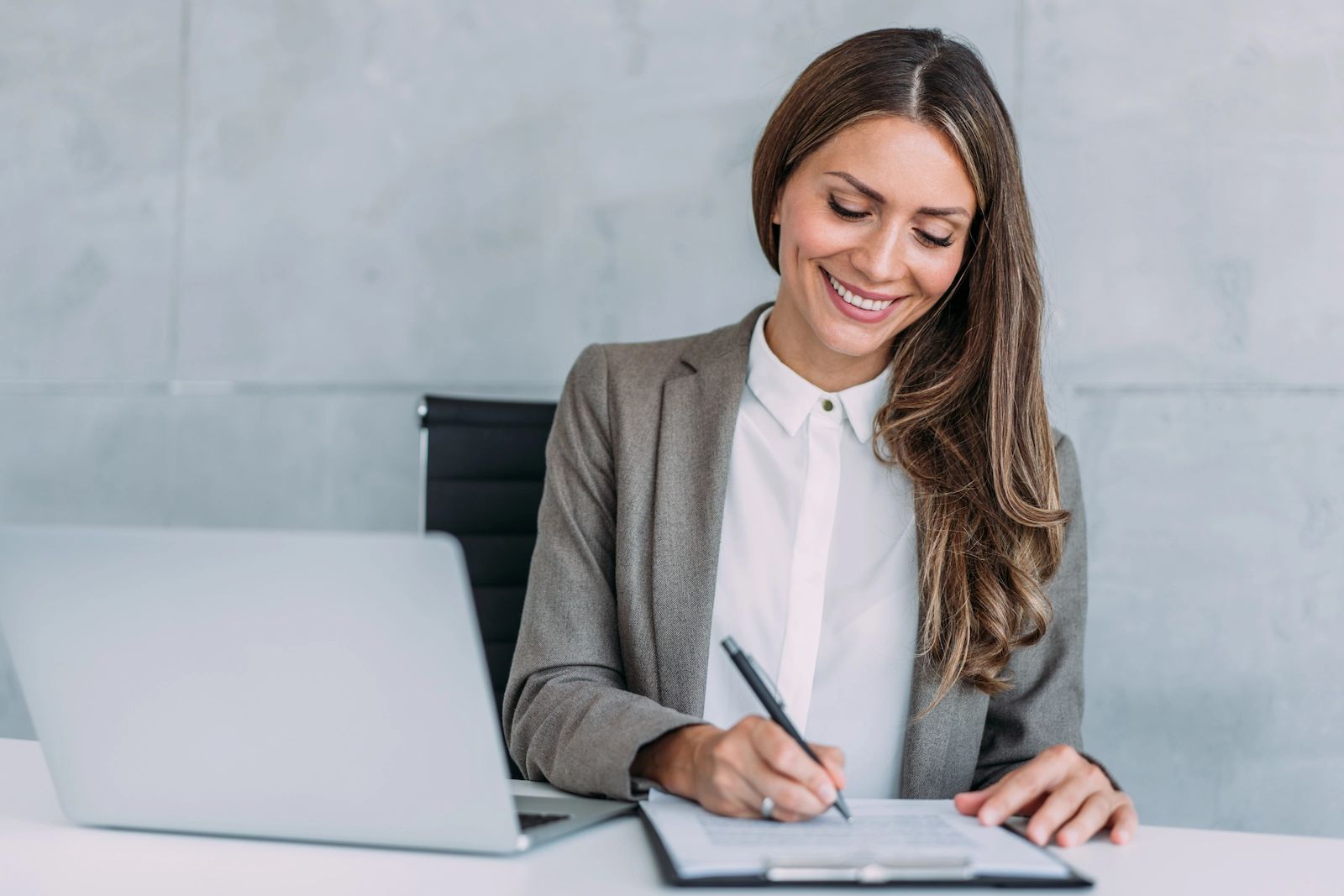 Eine lächelnde Geschäftsfrau im grauen Blazer unterschreibt Dokumente an ihrem Schreibtisch, neben ihr steht ein Laptop.