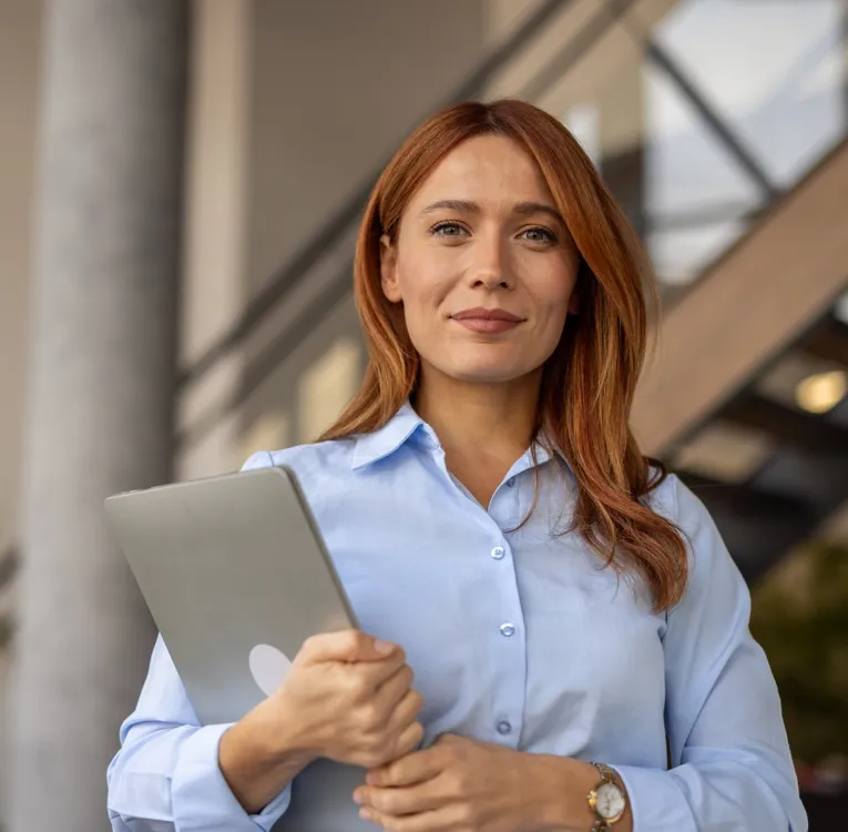 Eine selbstbewusste Geschäftsfrau mit roten Haaren in hellblauem Hemd hält einen Laptop, während sie in einem modernen Büro steht. Das Bild symbolisiert Führungsstärke, Technologie und effizientes Workforce Management.