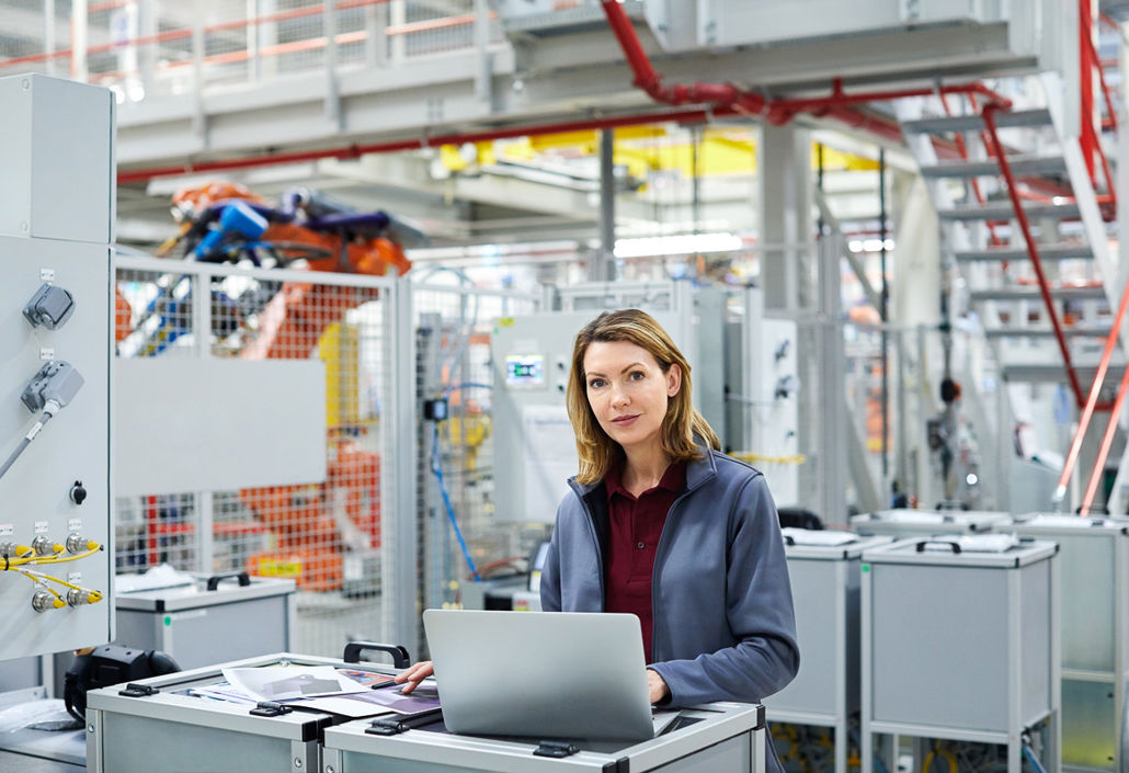 Eine selbstbewusste Ingenieurin arbeitet an einem Laptop in einer modernen Industrieanlage und überwacht automatisierte Produktionssysteme.