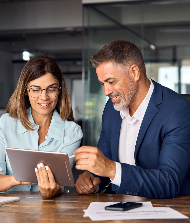 Zwei Geschäftsleute arbeiten gemeinsam in einem modernen Büro und betrachten Informationen auf einem Tablet. Die Frau trägt eine Brille und ein hellblaues Hemd, während der Mann im marineblauen Anzug lächelt und auf den Bildschirm zeigt. Ideal für Inhalte zu Workforce Management, Teamarbeit oder Unternehmensstrategie.