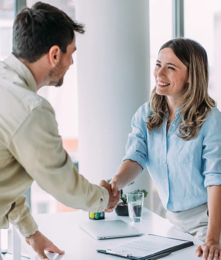 Lächelnde Geschäftsleute geben sich über einem Schreibtisch in einem hellen Büro die Hand, ein Symbol für erfolgreiche Zusammenarbeit, Onboarding und eine positive Arbeitskultur.